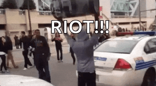 a man is standing in front of a police car holding a sign that says riot !!!