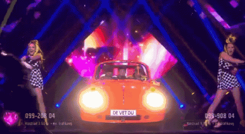 two women are dancing in front of an orange car with a license plate that says de vet du