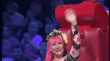 a woman with pink hair is sitting in a red chair and waving her hand .