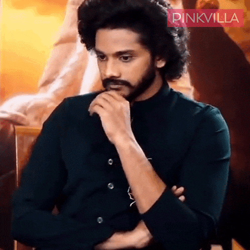 a man with curly hair and a beard is wearing a black shirt and a pinkvilla logo behind him