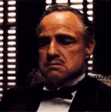 a man in a tuxedo and bow tie is sitting in front of a window with blinds .