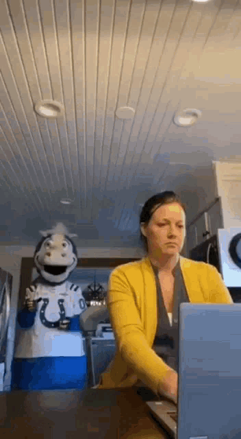 a woman is sitting at a table with a laptop and a mascot standing behind her .