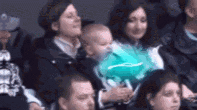 a group of people are sitting in a stadium watching a game and a baby is holding a green balloon .