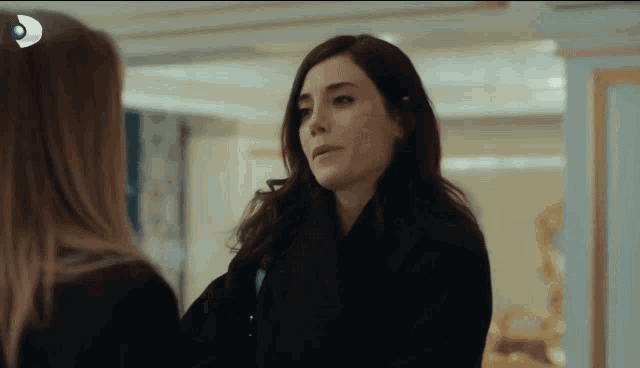 a woman in a black coat is talking to another woman in a room