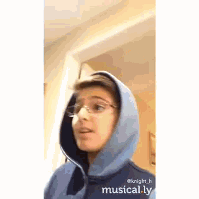 a young boy wearing a blue hoodie and glasses is standing in a room .