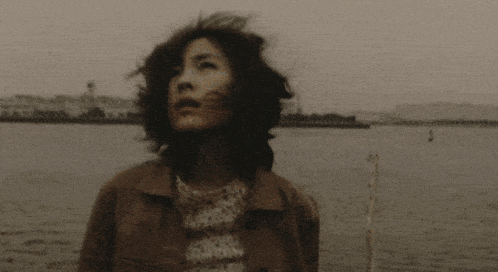 a woman stands in front of a body of water with her hair blowing in the wind