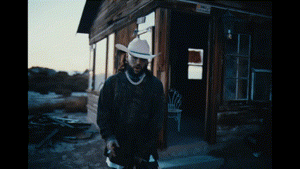 a man in a cowboy hat is standing in front of a wooden building holding a gun .