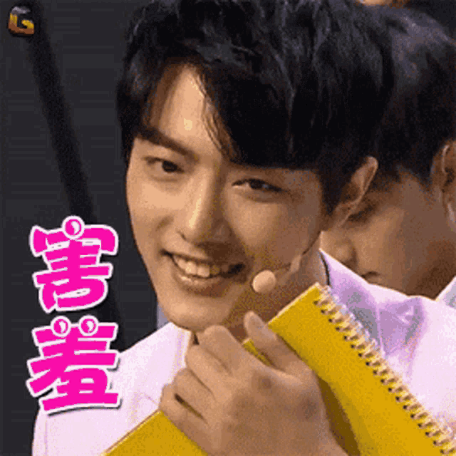 a man in a pink shirt is smiling and holding a yellow notebook