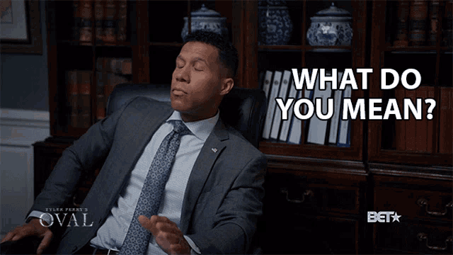 a man in a suit and tie is sitting in a chair with the words " what do you mean " behind him