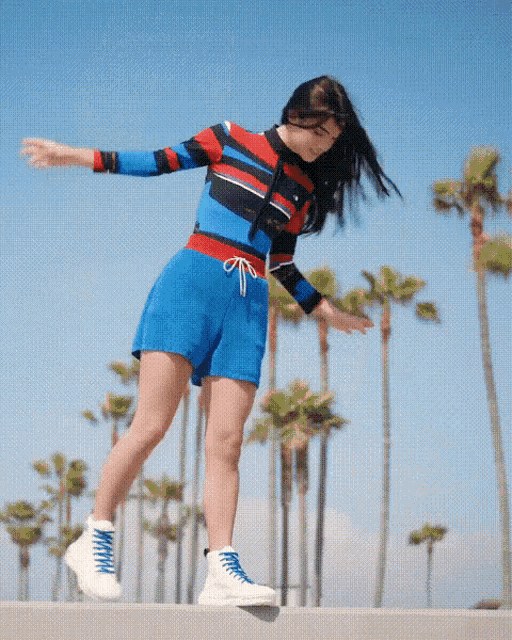 a woman in a striped shirt and blue shorts is standing on a ledge with her arms outstretched