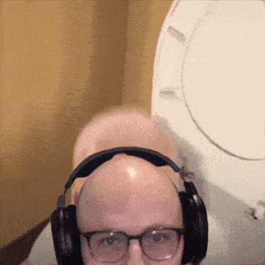 a bald man wearing glasses and headphones stands in front of a white toilet