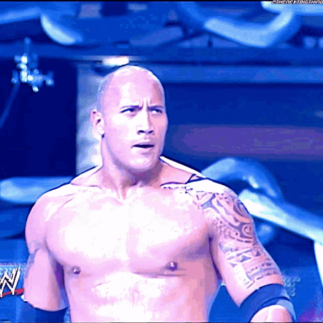 a shirtless wrestler with a tattoo on his arm stands in front of a blue background