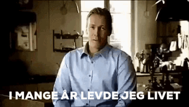 a man in a blue shirt is sitting in a kitchen and talking to the camera .