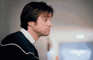 a man in a black sweater is leaning against a wall and making a sad face .
