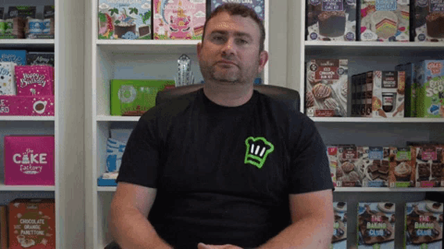 a man wearing a black shirt with a green chef 's hat on it sits in front of a cake factory shelf