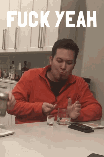 a man in a red jacket is sitting at a table with a shot glass and the words fuck yeah above him