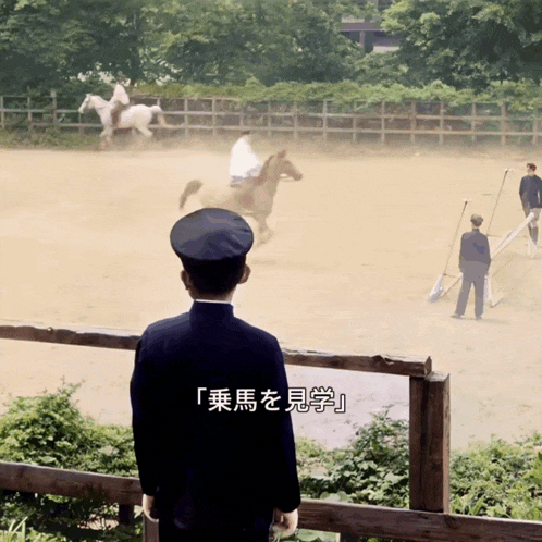 a man in a blue uniform stands in front of a fence watching horses in a stable