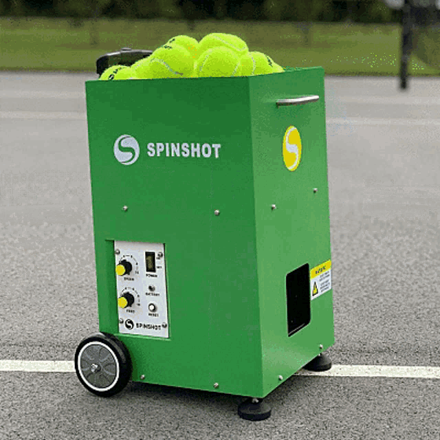 a green spinshot machine filled with tennis balls on a tennis court