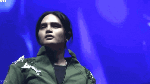 a woman wearing a green jacket stands in front of a blue background with the letters zky on it