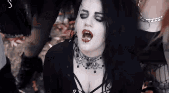 a woman with black hair and a choker is sitting on the ground with her mouth open and a group of people behind her .