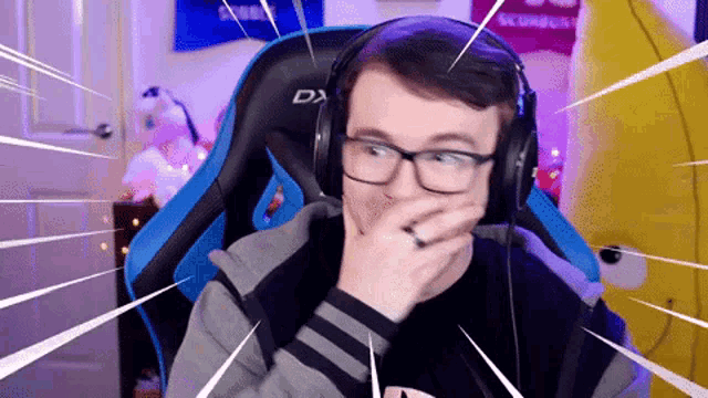 a man wearing glasses and headphones is sitting in a gaming chair and covering his mouth with his hand .