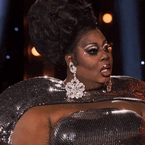 a drag queen wearing a silver dress and earrings looks to her left