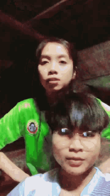 a girl wearing a green shirt with a beetle on it stands next to a boy wearing glasses