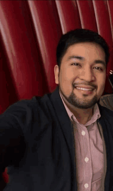 a man with a beard is smiling while taking a selfie in front of a red curtain .