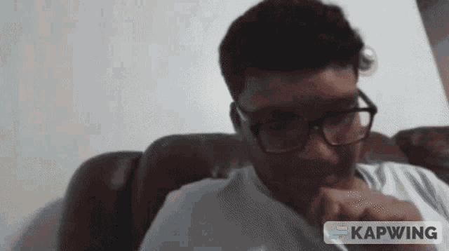 a man wearing glasses is sitting on a couch and the word kapwing is visible in the corner