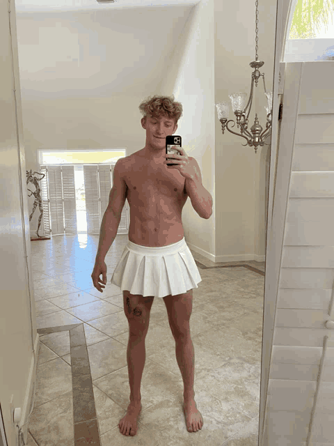 a shirtless man in a white pleated skirt takes a picture of himself in the mirror