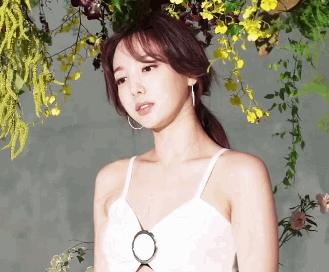a woman in a white tank top stands in front of a wall of flowers