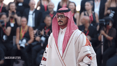 a man wearing a keffiyeh and glasses stands in front of a crowd of people with the number 1653181725 on the bottom right