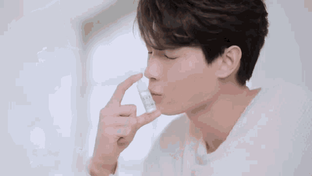 a man in a white sweater is holding a small bottle in his hand and spraying it into his nose .