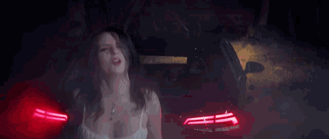 a woman in a white tank top is standing in front of a red car with smoke coming out of it .