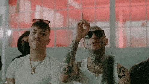 two men are standing next to each other in a room . one of the men is wearing sunglasses and a white tank top .