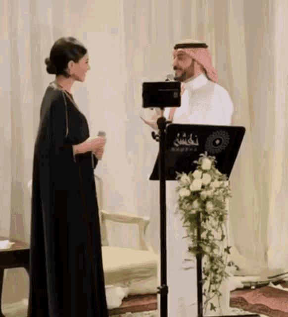 a man in a turban stands next to a woman holding a microphone in front of a podium with arabic writing