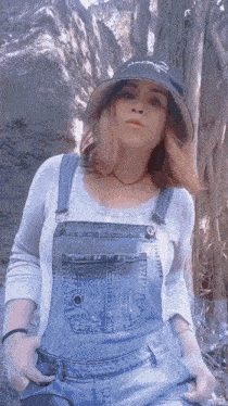 a woman wearing overalls and a hat has her hands in her pockets while standing in the woods