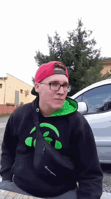 a young man wearing glasses and a red hat is standing in front of a car .