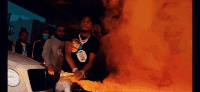 a group of men are standing in front of a burning car