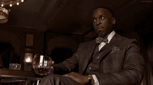a man in a suit and bow tie is sitting at a table with a glass of whiskey in his hand .
