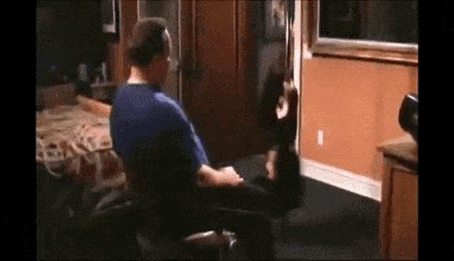 a man in a blue shirt is sitting in a chair in a living room with a woman standing behind him .