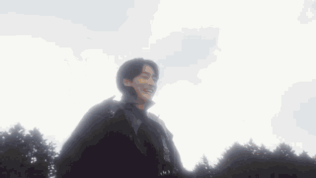 a man in a black jacket stands in front of a cloudy sky with trees in the background