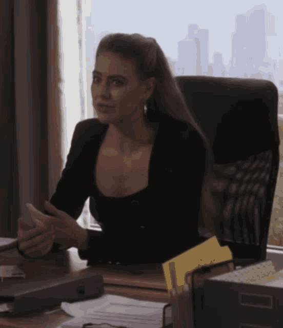 a woman in a black top sits at a desk in front of a window holding a cell phone