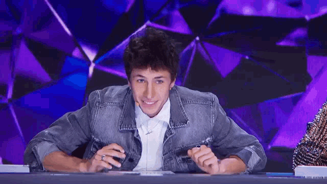 a man in a denim jacket is smiling while sitting at a table in front of a purple background .