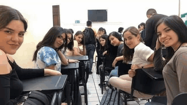 a group of young women are sitting at desks in a classroom and taking a selfie .