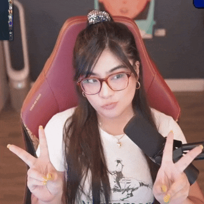 a woman wearing glasses and a ponytail is sitting in a chair with a microphone and making a peace sign .