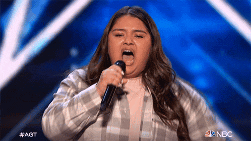 a girl is singing into a microphone on a stage with a nbc logo in the background