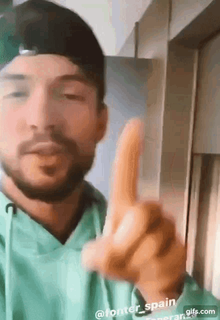 a man with a beard is wearing a green hoodie and a black hat and pointing his finger at the camera .