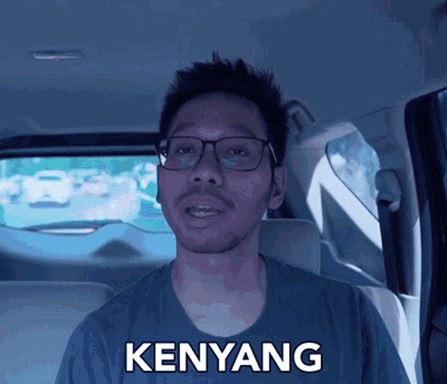 a man wearing glasses is sitting in the back seat of a car and says kenyang