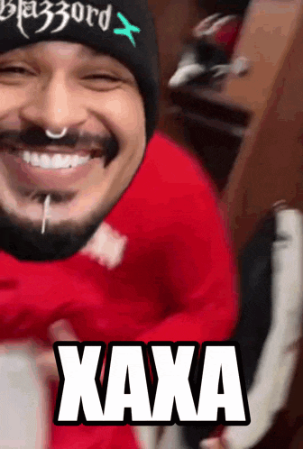 a man with a beard wearing a beanie and a red sweater says " kaxa " in white letters